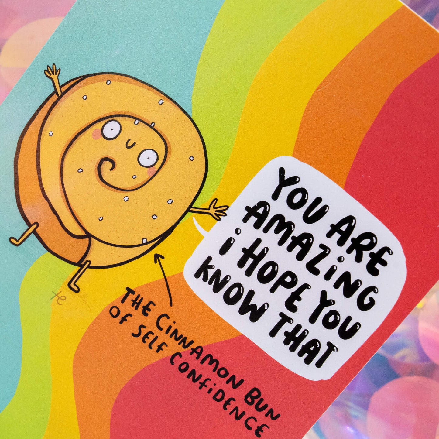 Close-up of The Cinnamon Bun of Self Confidence postcard by Happiness Enchanters, showing the happy cinnamon bun character and bold positive message “You are amazing I hope you know that,” surrounded by rainbow colours and glossy silk finish detail.