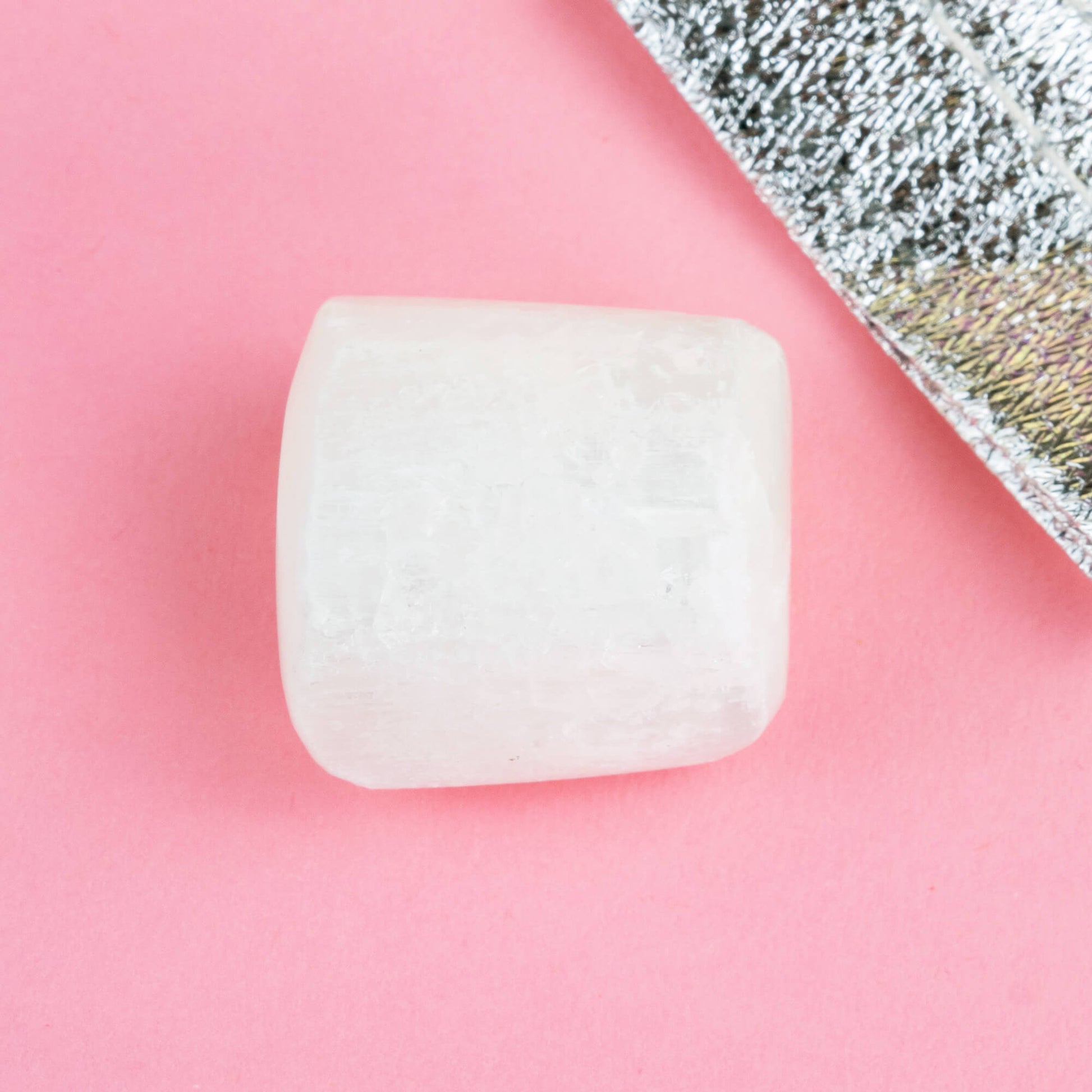 Happiness Enchanters selenite crystal on a pink backdrop with metallic silver bag laid next to it