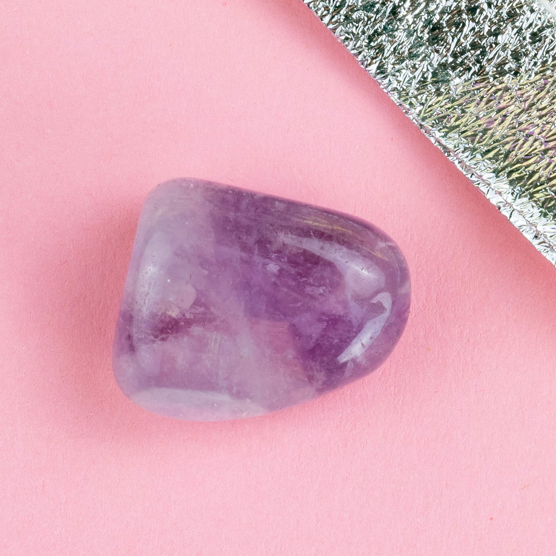 Happiness Enchanters amethyst crystal on a pink backdrop with metalic silver bag laid next to it