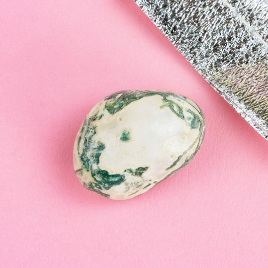 Happiness Enchanters tree agate crystal on a pink backdrop with metallic silver bag laid next to it