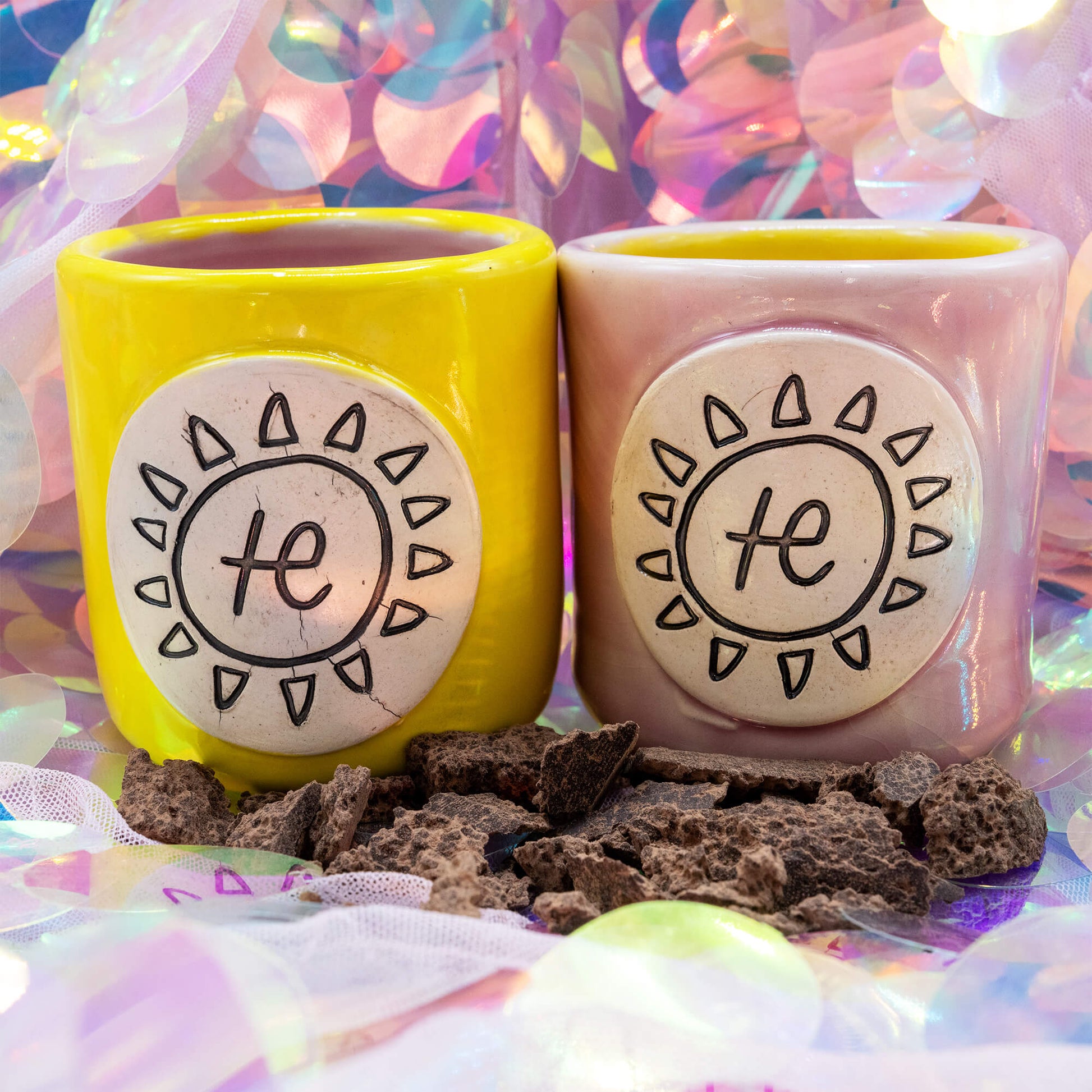 Pair of yellow and pink handmade ceramic cacao cups by Happiness Enchanters with carved sun symbols, displayed on a shimmering pastel background surrounded by pieces of raw cacao.