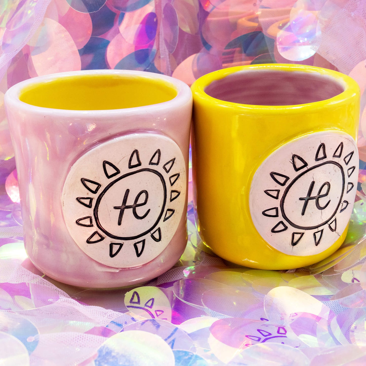 Pink and yellow handmade cacao mugs by Happiness Enchanters with white sun plaque designs, side by side on a holographic background.