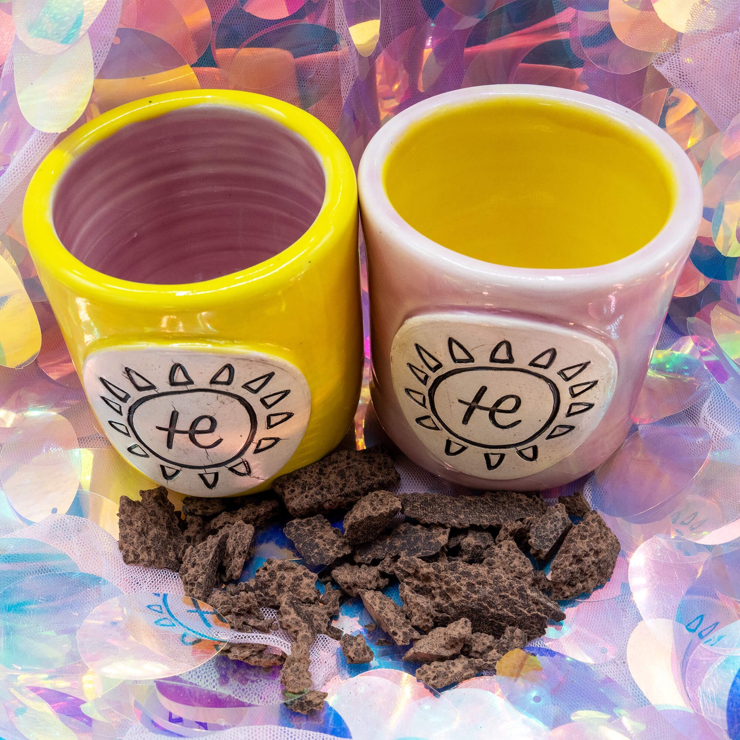 Close-up of Happiness Enchanters handmade pink and yellow cacao cups surrounded by pieces of raw cacao on a pastel iridescent surface.