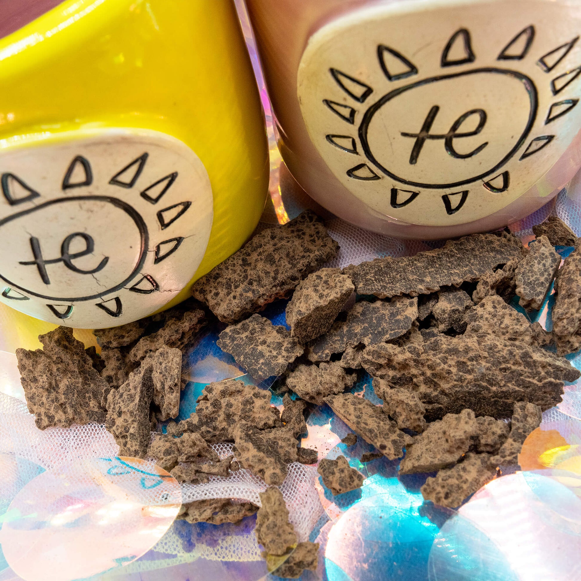 Close-up of Happiness Enchanters handmade pink and yellow ceramic cacao mugs with white sun plaques and chunks of raw cacao on an iridescent fabric surface.