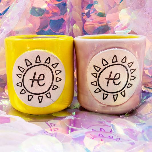 Pair of yellow and pink Happiness Enchanters handmade cacao cups with hand-carved sun emblems, placed together on an iridescent background.