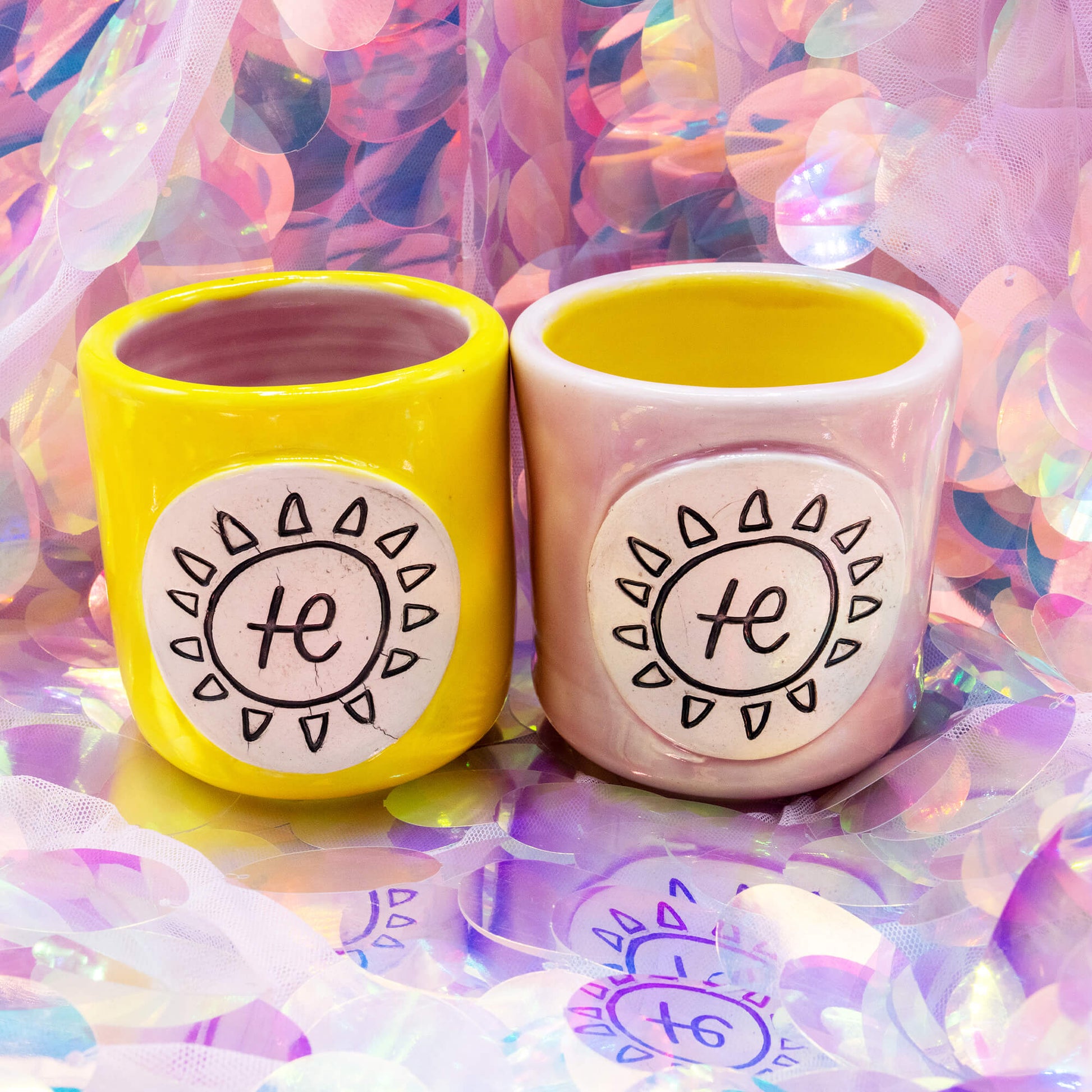 Front view of yellow and pink handmade Happiness Enchanters mugs with sun and symbol design plaques.