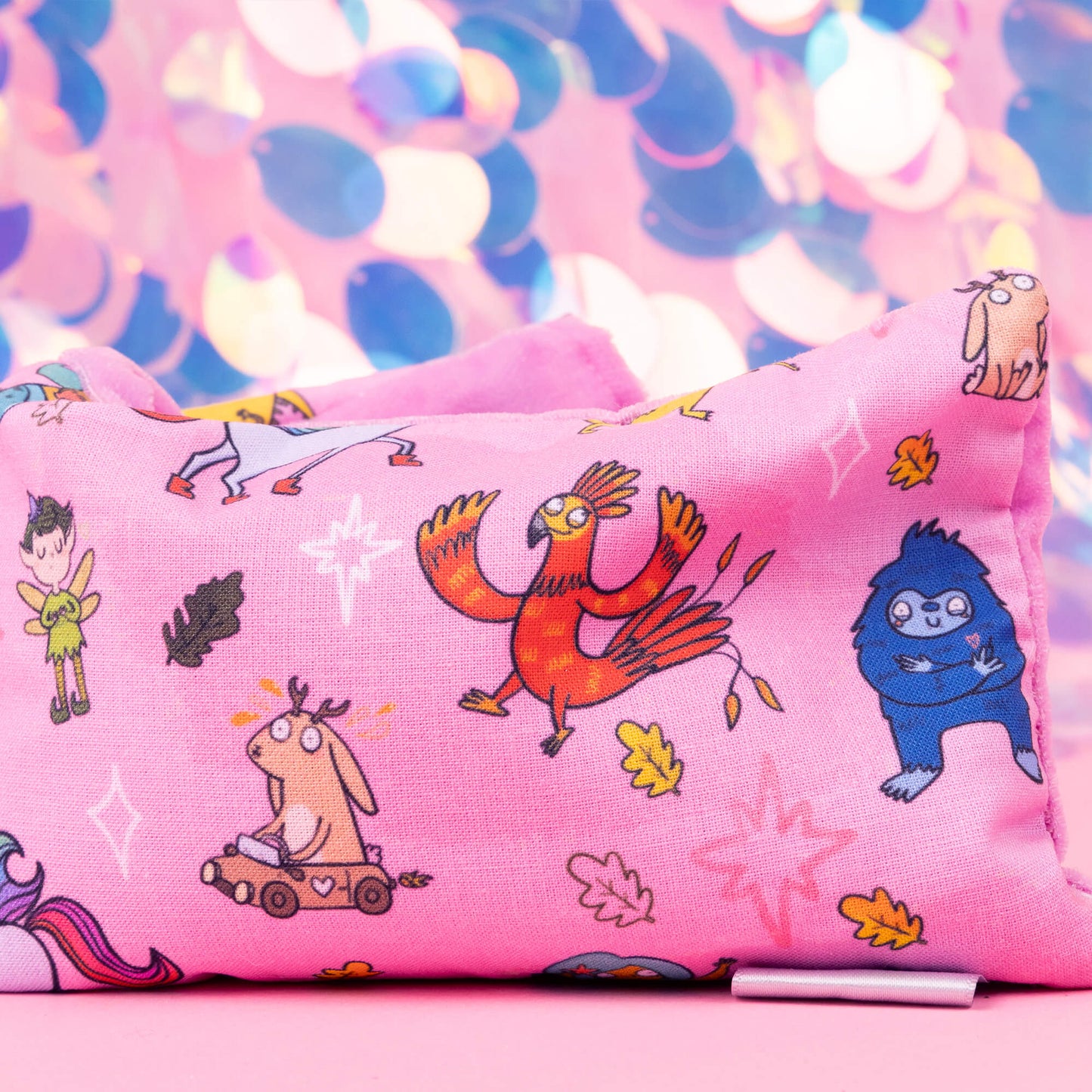 A detailed view of the Happiness Enchanters x Dawn's Sewing Room Eye Bags Relaxing Eye Pillow showcasing a pink fabric cover decorated with various charming characters and elements illustrated by Katie Abey. The colourful and playful design features fairies, animals, and mythical creatures, set against a pastel bokeh background that adds a magical touch