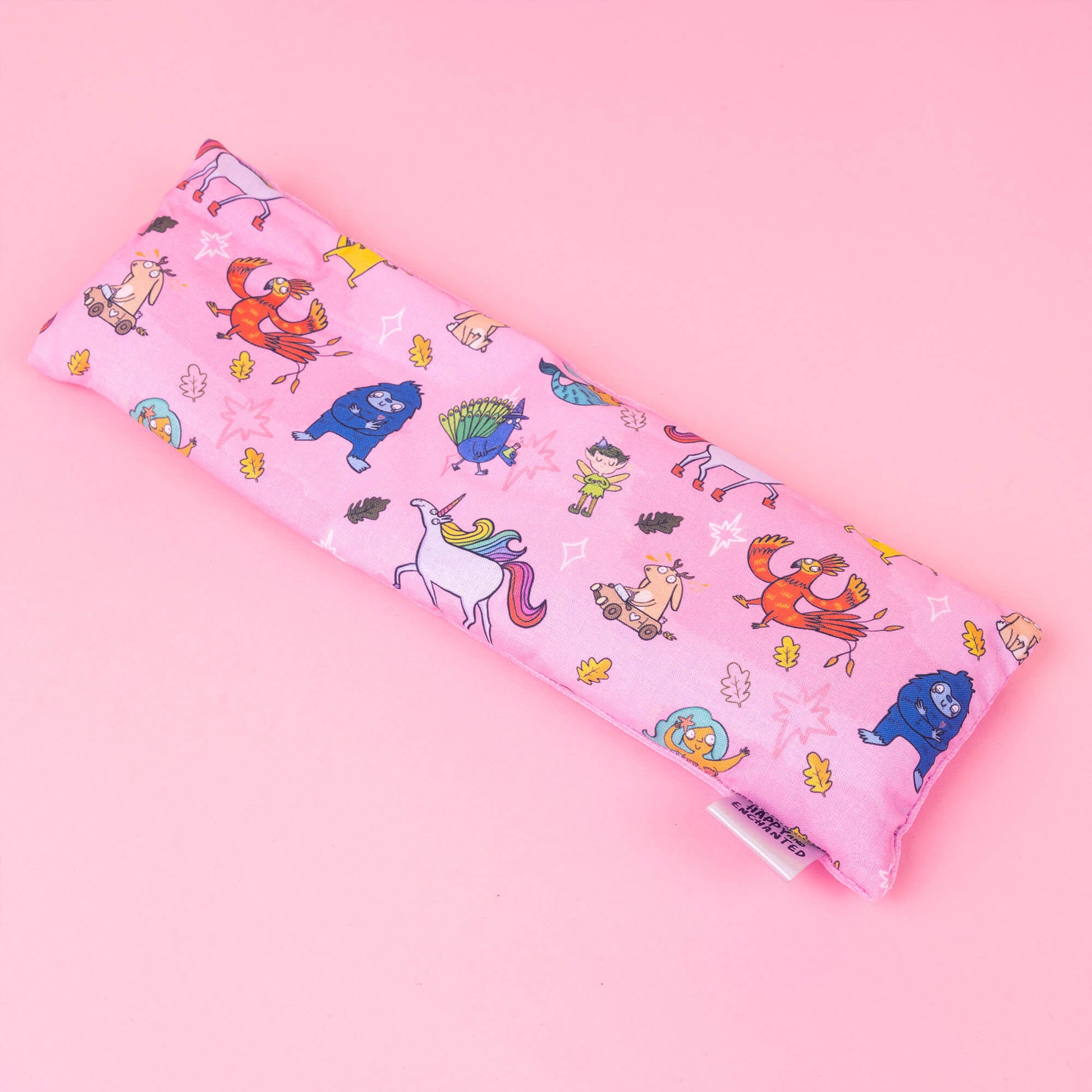 A full-length image of the Happiness Enchanters x Dawn's Sewing Room Eye Bags Relaxing Eye Pillow, displaying the entirety of the pink fabric cover illustrated with Katie Abey's whimsical characters. The design includes a mix of fairies, animals, and mythical creatures, all set against a solid pink background, highlighting the vibrant and playful artwork.