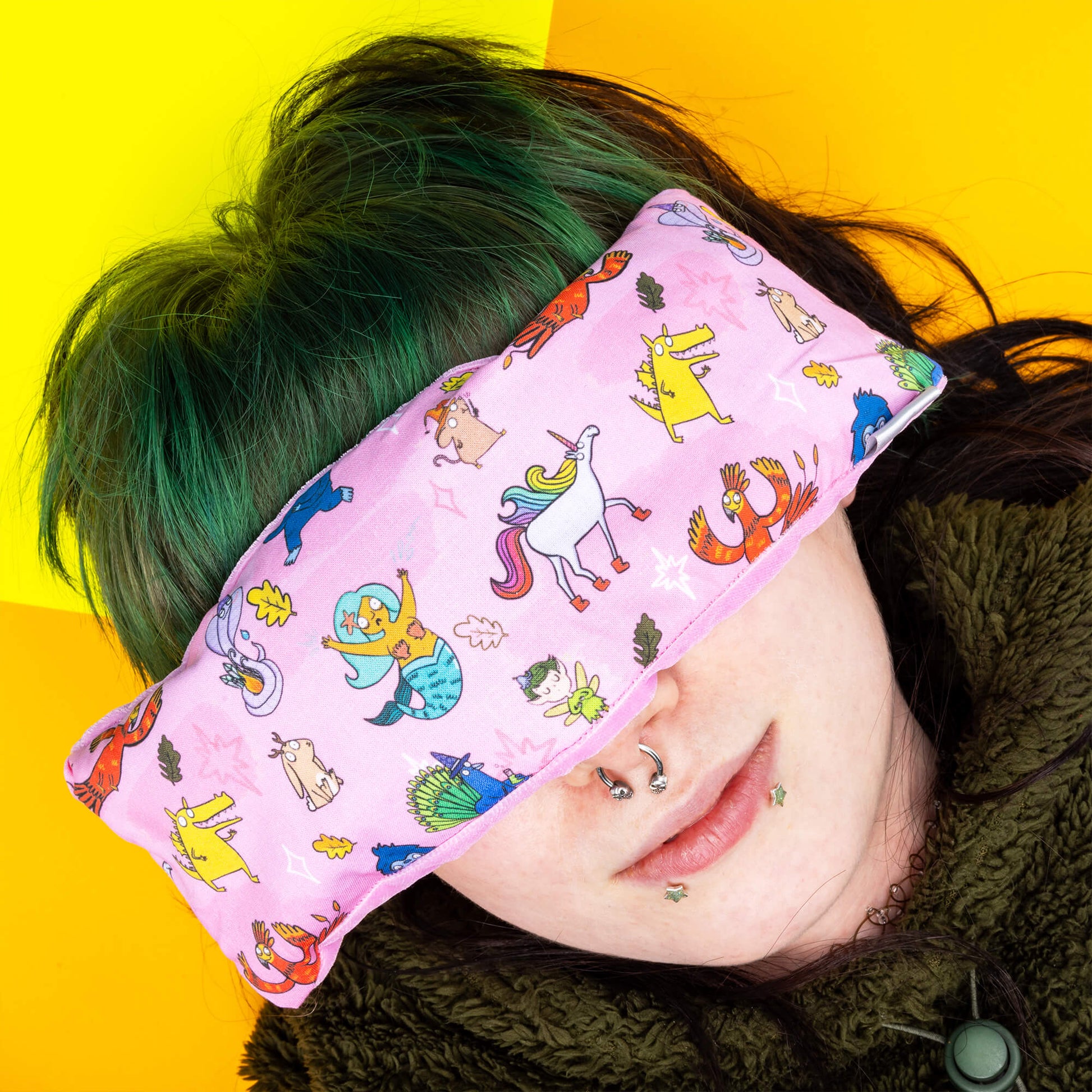 A person with green hair is wearing Happiness Enchanters x Dawn's Sewing Room Eye Bags Relaxing Eye Pillow. The eye mask features a pink fabric cover with delightful illustrations by Katie Abey, including various colourful characters such as unicorns, fairies, and animals. The person lies against a contrasting yellow background, showcasing the mask's design and fit