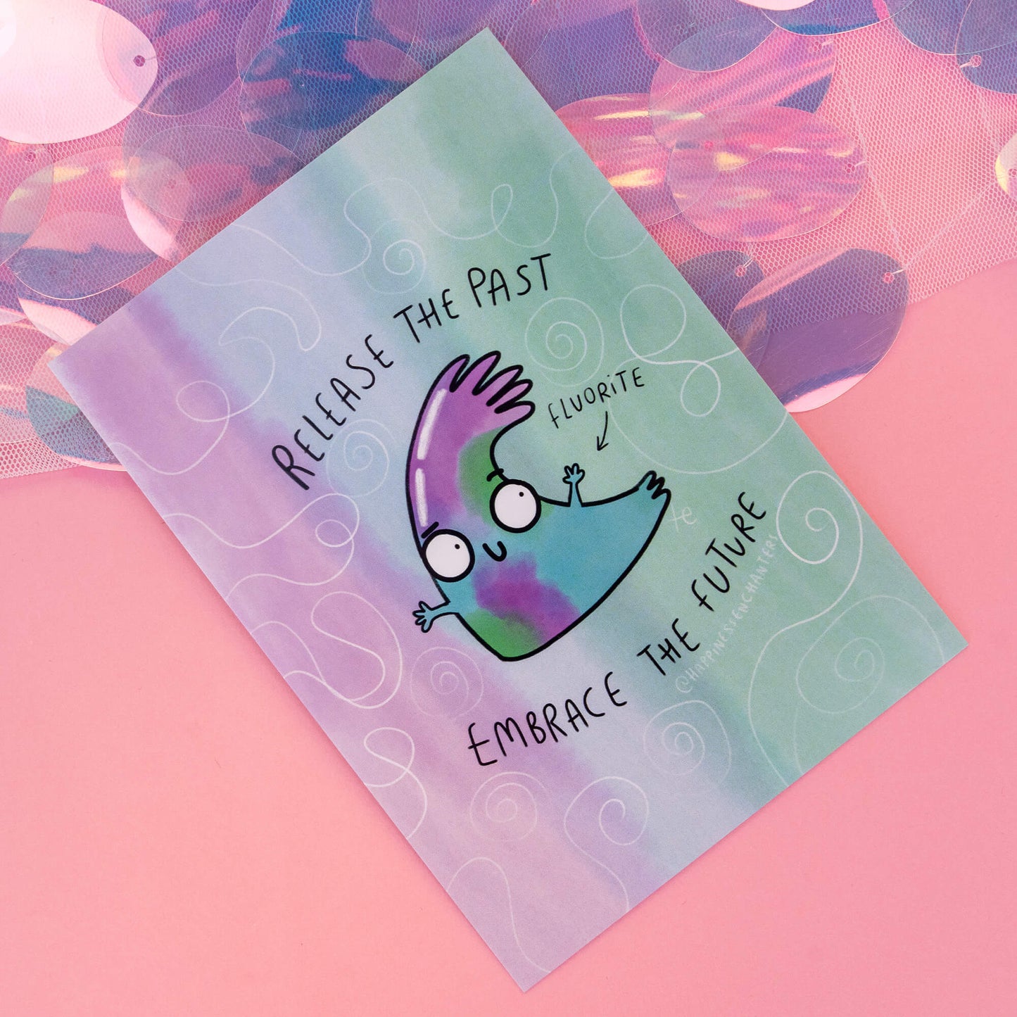 A close-up view of a Fluorite-themed postcard by 'Happiness Enchanters', displaying a charming cartoon Fluorite crystal with the phrase 'Release the past, embrace the future'. The background is a soft blend of pastel colours with swirling patterns, capturing the essence of creativity and enchantment.