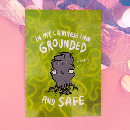 A close-up view of a Hematite-themed postcard by 'Happiness Enchanters', displaying a charming cartoon Hematite crystal with the phrase 'In my calmness I am grounded and safe'. The background is a vibrant green with swirling patterns, capturing the essence of creativity and enchantment.