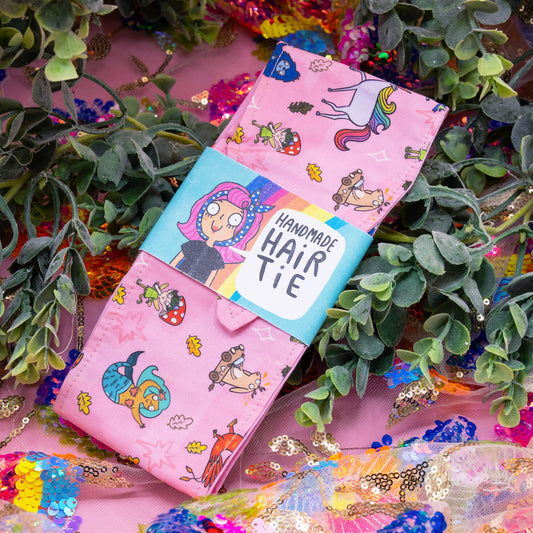 Handmade Word Spells Hair Tie by Happiness Enchanters, featuring a whimsical pink design with magical creatures and retro vibes. Designed by Katie Abey and Angela Sandland, made from cotton with a reversible toning backing. Perfect for adding charm to hairstyles or taming bad hair days. Displayed among colourful sequins and green foliage.