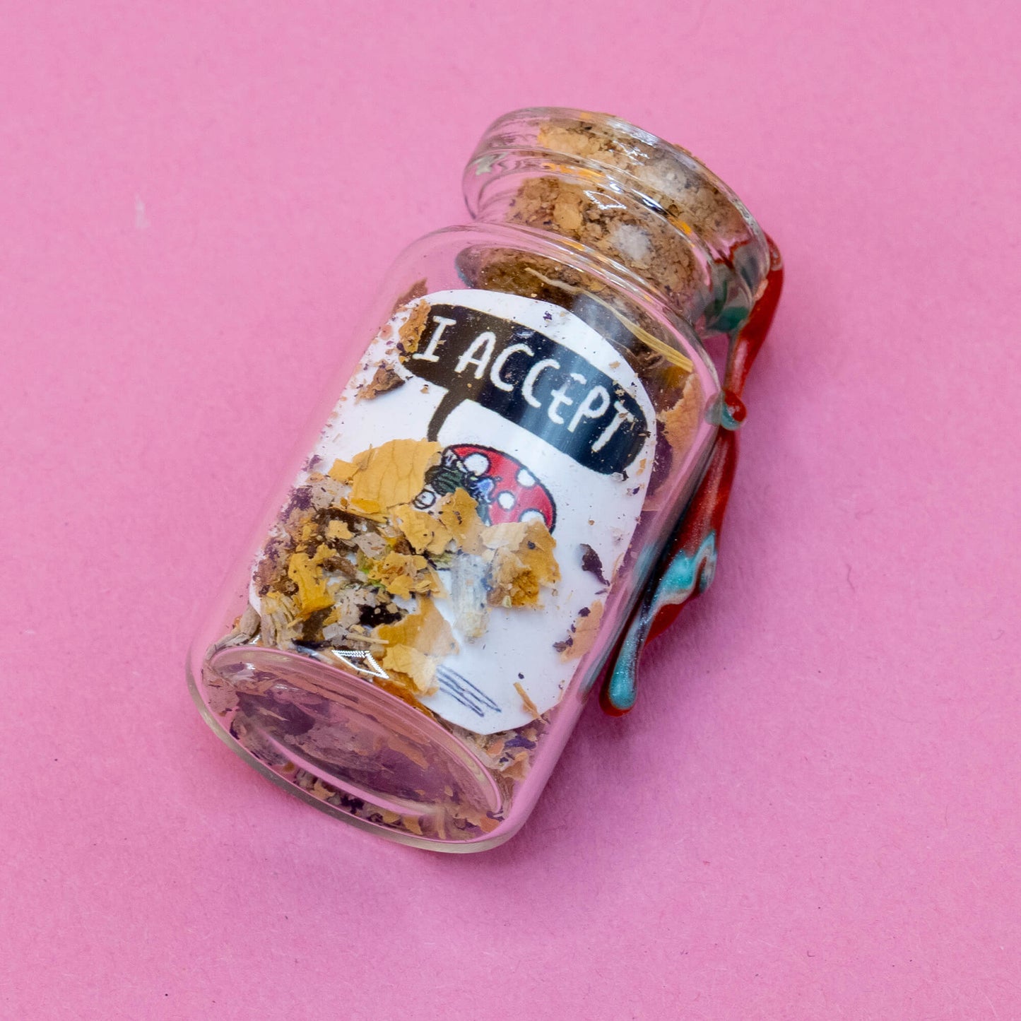 A small Pixie spell bottle lying on a pink surface, sealed with swirling red and white wax. The clear glass bottle is filled with dried herbs, petals, and magical ingredients. The label shows a red mushroom-capped Pixie character saying “I ACCEPT.” A handcrafted item from the Word Spells collection by Katie Abey & Angela Sandland.