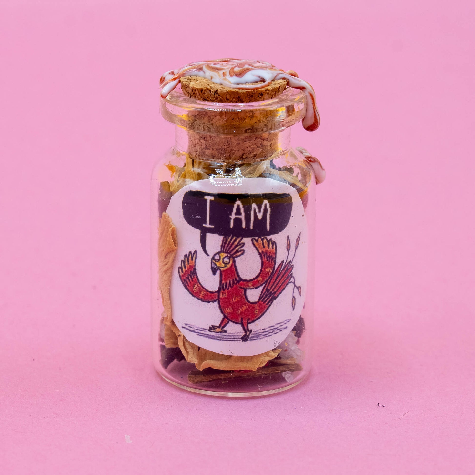 A small Phoenix spell bottle standing upright on a pink background. The clear glass jar is sealed with red and white swirled wax and a cork. The label features a vibrant red phoenix with wings spread, saying “I AM.” Inside is a mix of dried flowers, herbs, and crystals. A handmade creation from the Word Spells collection by Katie Abey & Angela Sandland.