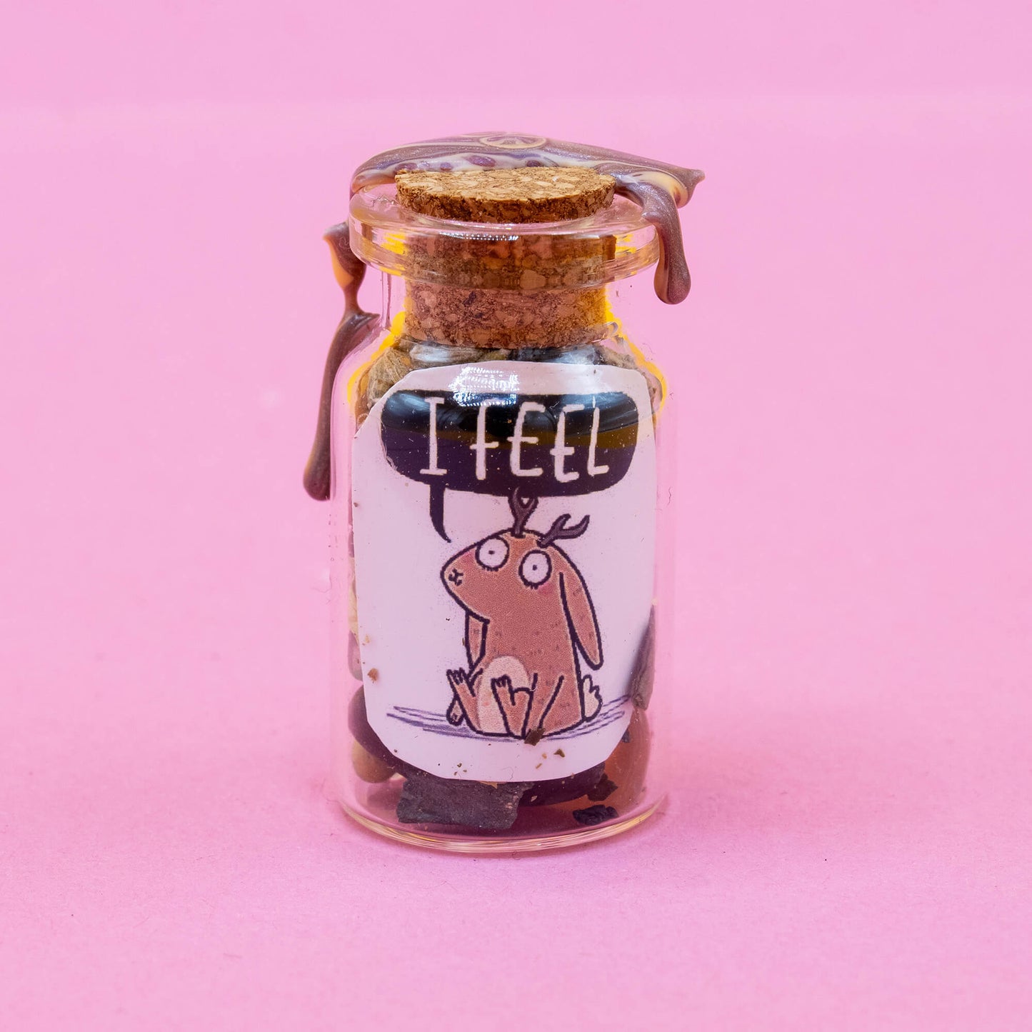 A jackalope spell bottle standing upright on a soft pink backdrop. Filled with magical herbs and crystals, and sealed with earthy-toned wax. Label features a cute jackalope saying “I FEEL.” A spiritual tool from the Word Spells range by Happiness Enchanters, crafted for self-connection, emotional expression, and ritual practice.