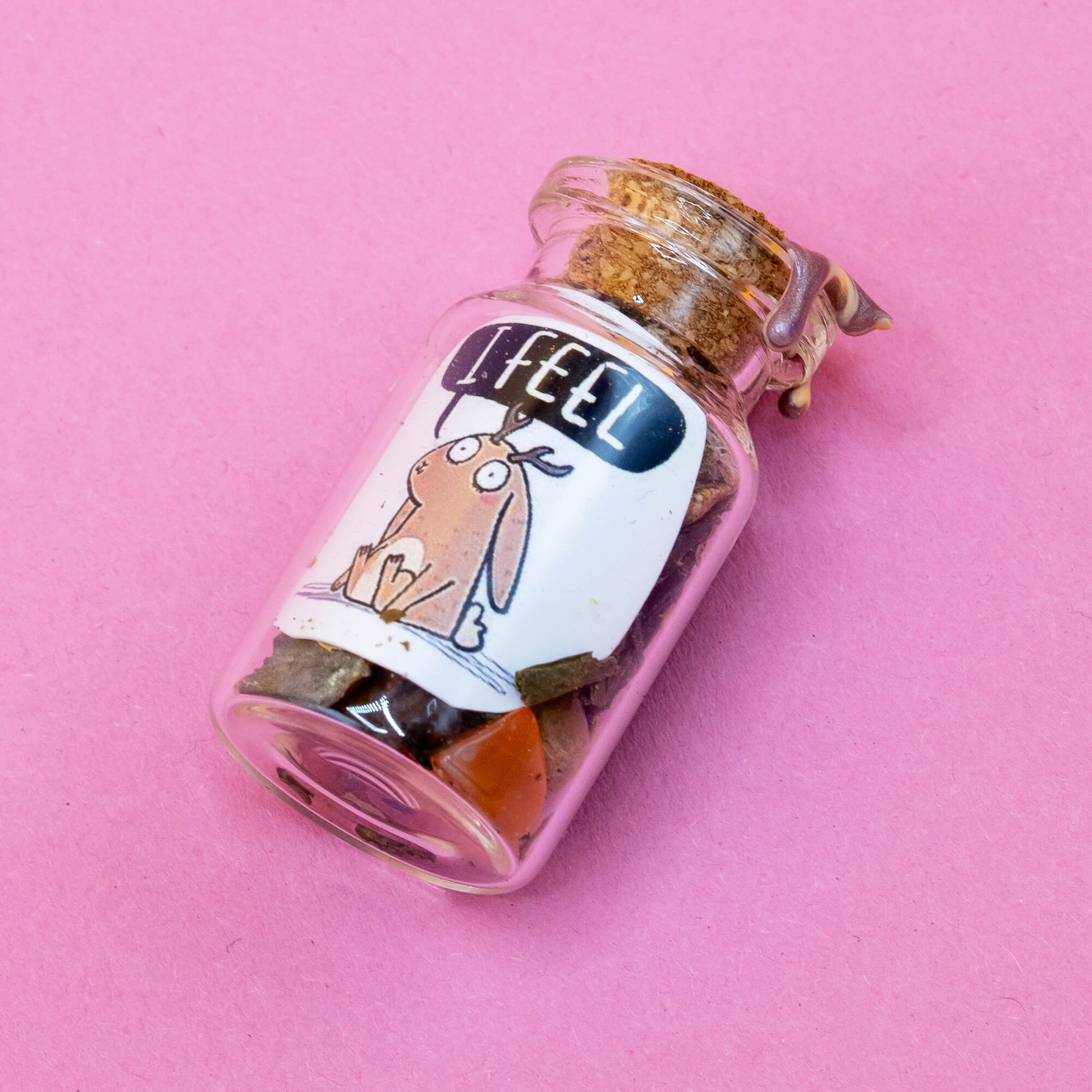 A small spell bottle lying on a pink background, filled with crystals and dried botanicals. Sealed with cork and marbled brown wax. Label shows a jackalope with a gentle expression and the words “I FEEL.” Created by Happiness Enchanters to inspire mindfulness and intention, part of the Word Spells series by Katie Abey & Angela Sandland.