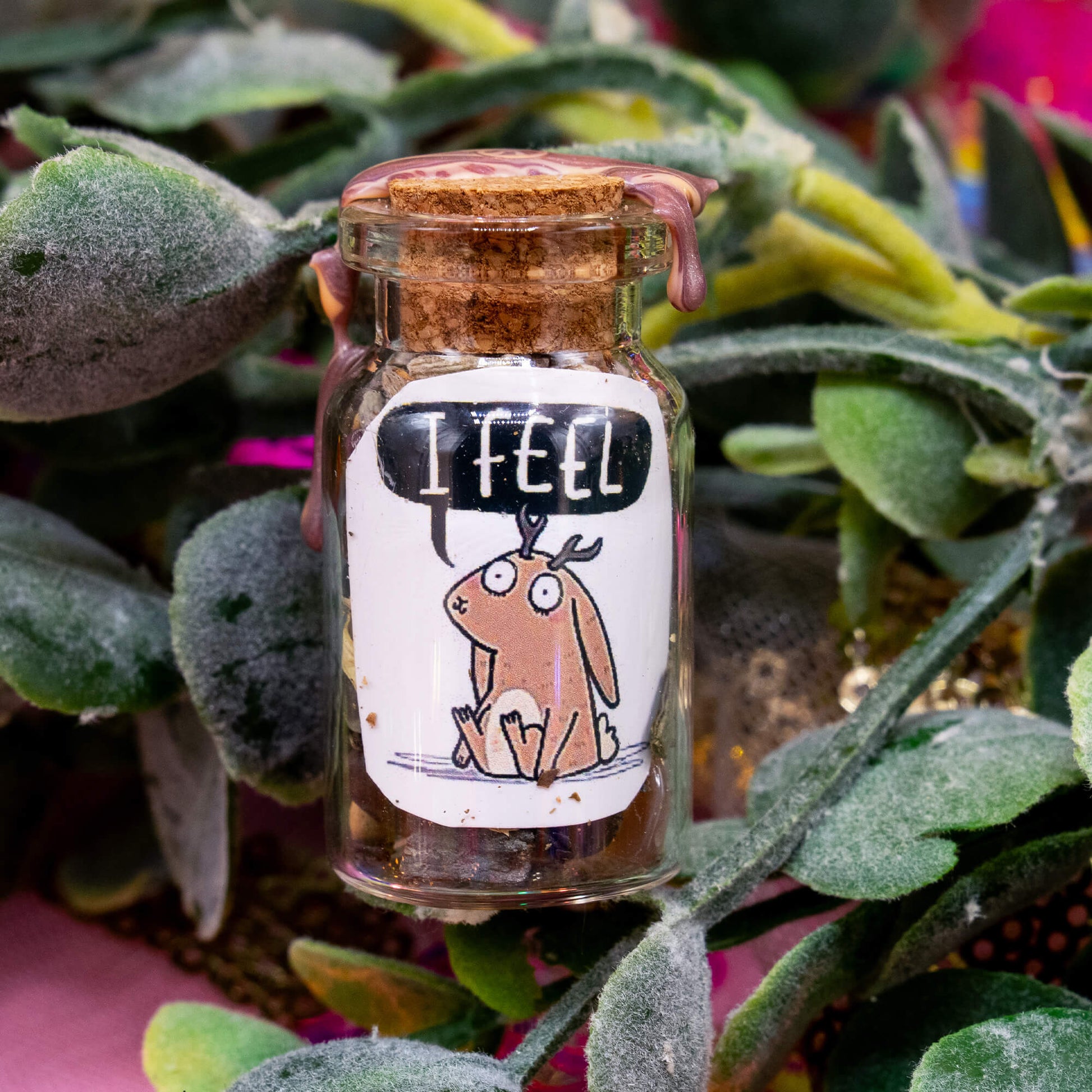 A handmade glass spell bottle nestled in green foliage, sealed with a cork and marbled brown wax. The front label features a sweet jackalope illustration with a speech bubble saying “I FEEL.” Filled with herbs, crystals, and magical materials. From the Word Spells collection by Katie Abey & Angela Sandland, designed to support emotional awareness and healing.