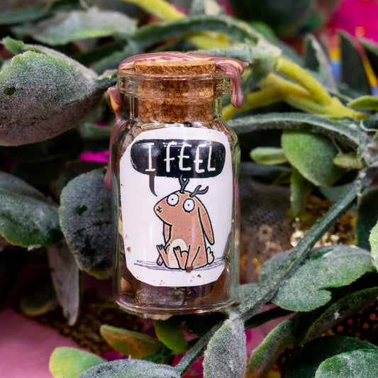 A handmade glass spell bottle nestled in green foliage, sealed with a cork and marbled brown wax. The front label features a sweet jackalope illustration with a speech bubble saying “I FEEL.” Filled with herbs, crystals, and magical materials. From the Word Spells collection by Katie Abey & Angela Sandland, designed to support emotional awareness and healing.