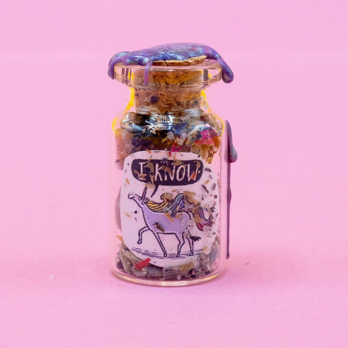 Glass unicorn spell bottle standing upright on a pastel pink backdrop, filled with a unique blend of herbs and crystals. Sealed with shimmering purple wax and cork. Features a magical “I KNOW” unicorn character label. A spiritual item from the Word Spells range by Happiness Enchanters, crafted by Katie Abey & Angela Sandland.
