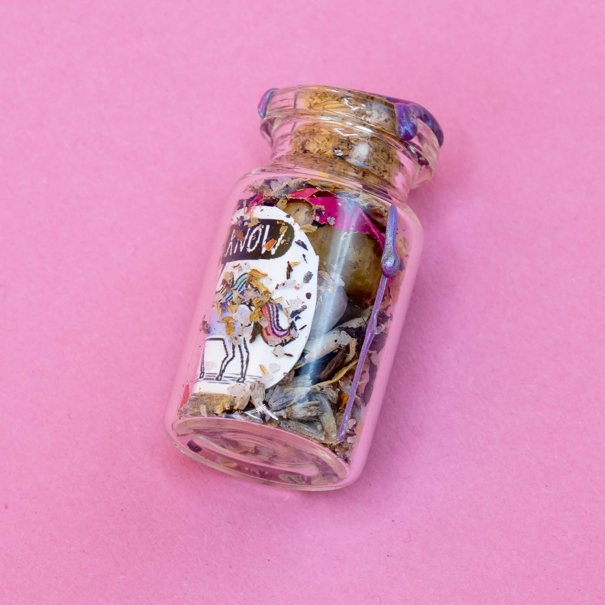 A handmade unicorn spell bottle lying on a pink background. The glass jar is filled with magical herbs and crystals, sealed with purple wax. A mystical unicorn with rainbow mane and “I KNOW” speech bubble is illustrated on the label. Part of the Word Spells range by Katie Abey & Angela Sandland for Happiness Enchanters.