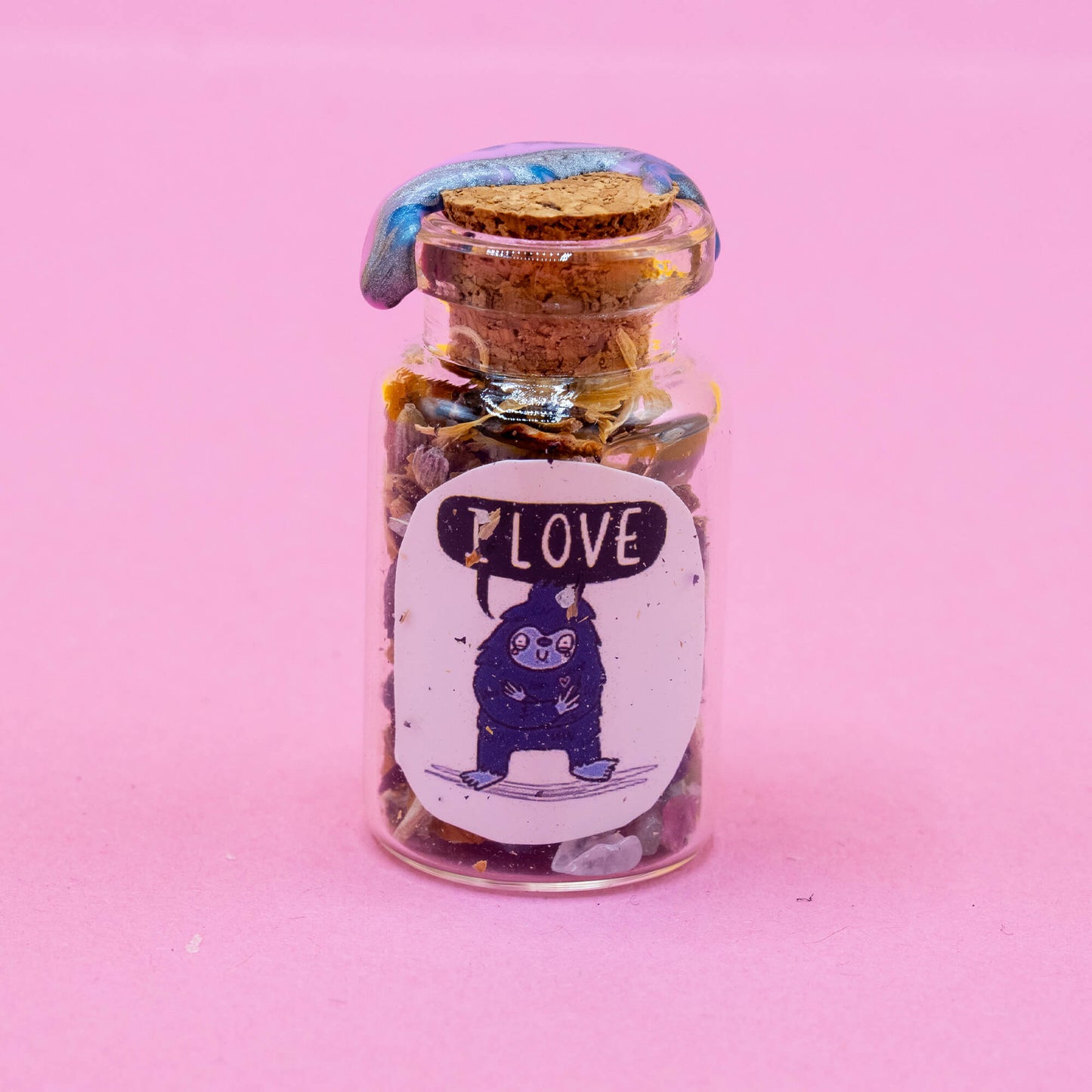 A Bigfoot-themed spell bottle standing upright on a pastel pink background. The glass jar is filled with dried flowers, crystals, and magical ingredients, sealed with cork and shimmering blue wax. The illustrated label shows a happy Bigfoot character saying “I LOVE.” Part of the Word Spells collection by Happiness Enchanters, created by Katie Abey & Angela Sandland.