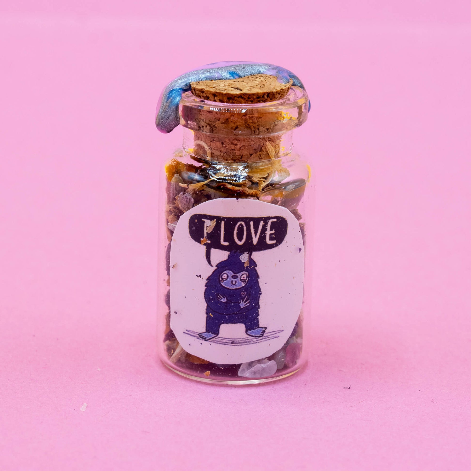 A Bigfoot-themed spell bottle standing upright on a pastel pink background. The glass jar is filled with dried flowers, crystals, and magical ingredients, sealed with cork and shimmering blue wax. The illustrated label shows a happy Bigfoot character saying “I LOVE.” Part of the Word Spells collection by Happiness Enchanters, created by Katie Abey & Angela Sandland.