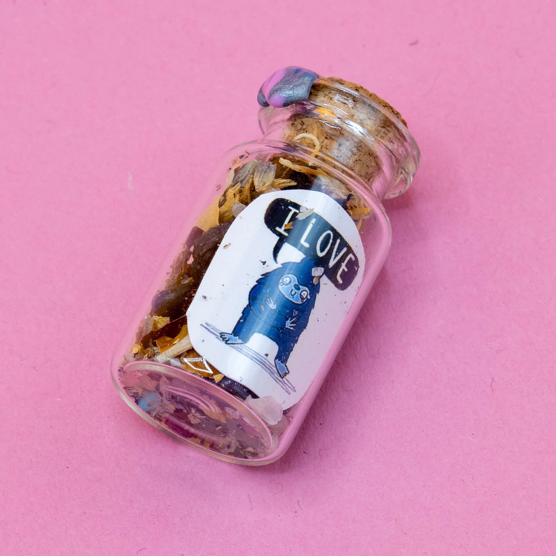 A small spell bottle lying on a pink surface, sealed with cork and blue wax. Inside are herbs, crystals, and love-filled magic. Label displays a friendly Bigfoot character saying “I LOVE.” One of the handmade Word Spell bottles by Katie Abey & Angela Sandland for intention-setting and heart-opening rituals.
