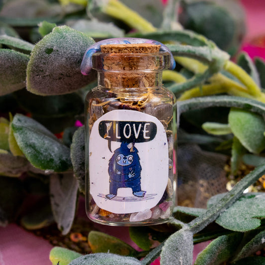 A handcrafted spell bottle nestled among green foliage and pink hues. The bottle contains a mix of herbs and crystals, sealed with iridescent wax. The label features a blue Bigfoot character with a sweet smile and a speech bubble that reads “I LOVE.” A spiritual tool from the Word Spells range by Happiness Enchanters.