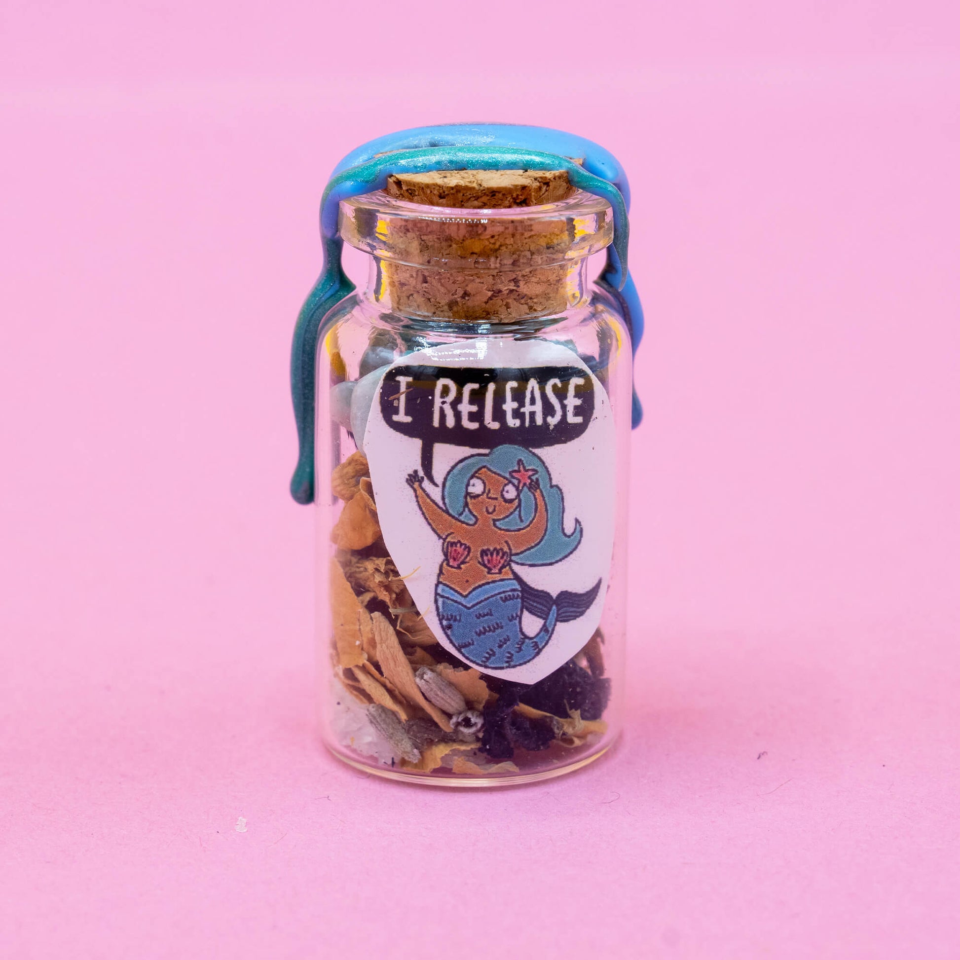 Glass spell bottle standing upright on a pink background, featuring a mermaid character with the affirmation “I RELEASE.” The bottle is filled with magical herbs and crystal trinkets, sealed with wax. From the Word Spells series by Katie Abey & Angela Sandland, created to support intention setting and emotional healing.