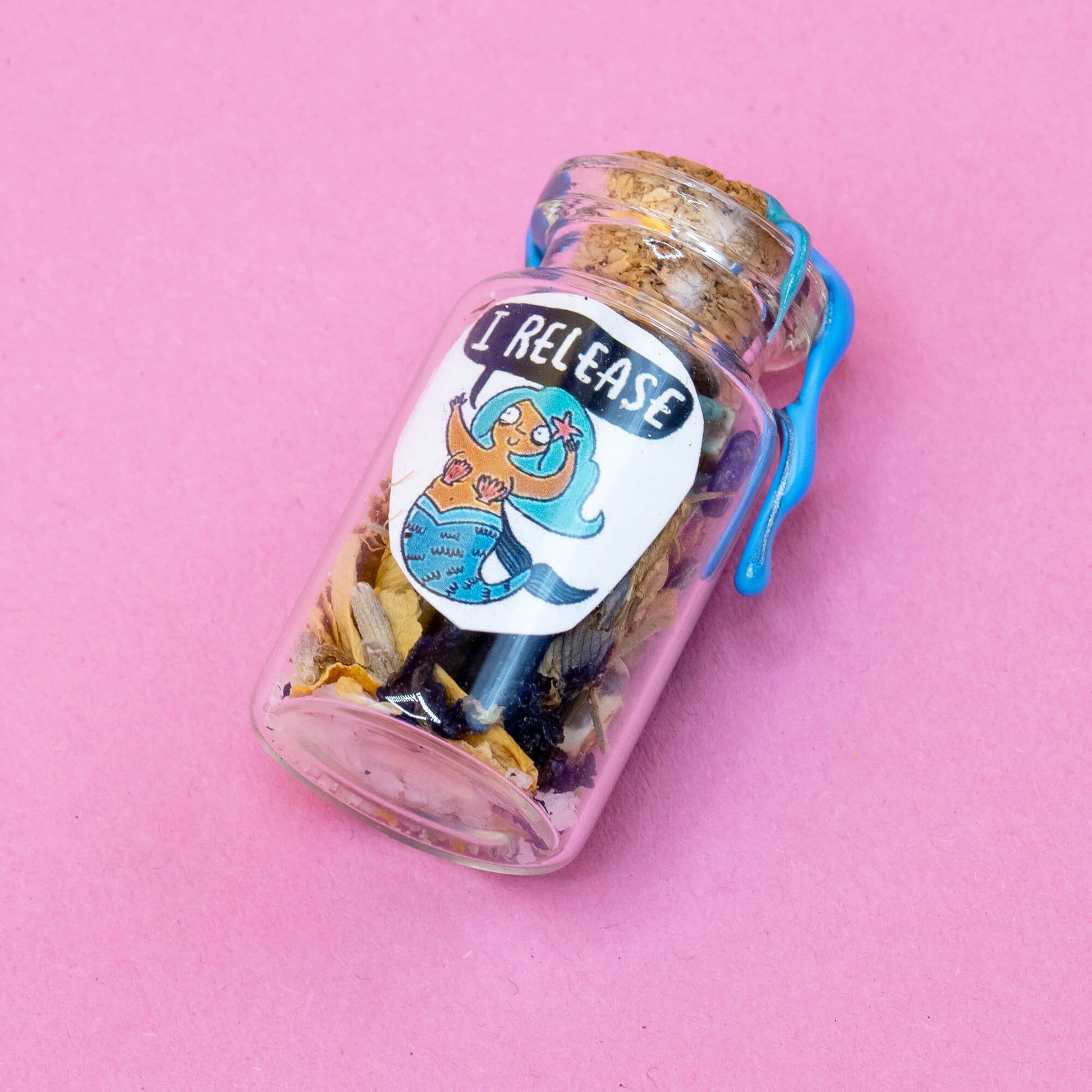 A small handmade spell bottle lying on a pink surface, filled with dried herbs, flowers, and crystals. Sealed with a cork and blue wax. The front label features a cheerful mermaid with turquoise hair and a speech bubble saying “I RELEASE.” Part of the Word Spells range by Happiness Enchanters, created by Katie Abey & Angela Sandland for spiritual and meditation use.