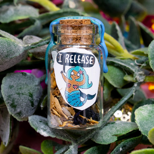 A close-up of a mermaid spell bottle nestled in soft green leaves. The glass jar contains a unique mix of herbs and crystals, sealed with blue wax. The illustrated label shows a mermaid character saying “I RELEASE.” A magical item from the Word Spells collection by Happiness Enchanters for meditation and personal rituals.