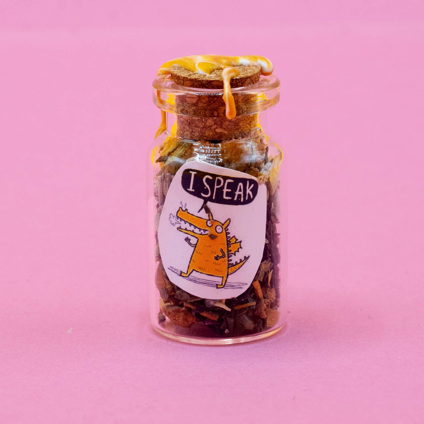 A handmade glass spell bottle standing upright on a pink background, sealed with yellow wax and a cork. The label shows a cheerful orange dragon breathing a tiny puff of smoke and saying “I SPEAK.” The jar is filled with herbs, petals, and magical materials. Part of the Word Spells collection by Katie Abey & Angela Sandland, supporting confident self-expression.
