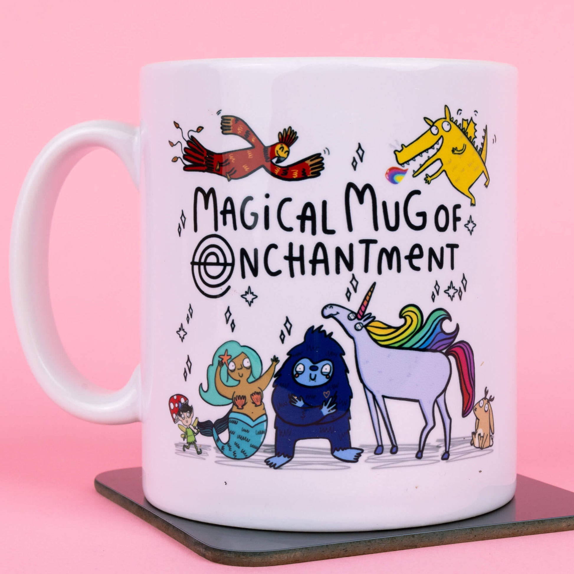 A white mug featuring whimsical characters designed by Katie Abey, including a vibrant rainbow unicorn, a blue creature, a mermaid, and a cheerful bird. Text reads "Magical Mug of Enchantment," surrounded by magical sparkles, against a pink background. Created by Happiness Enchanters, blending art and enchantment.