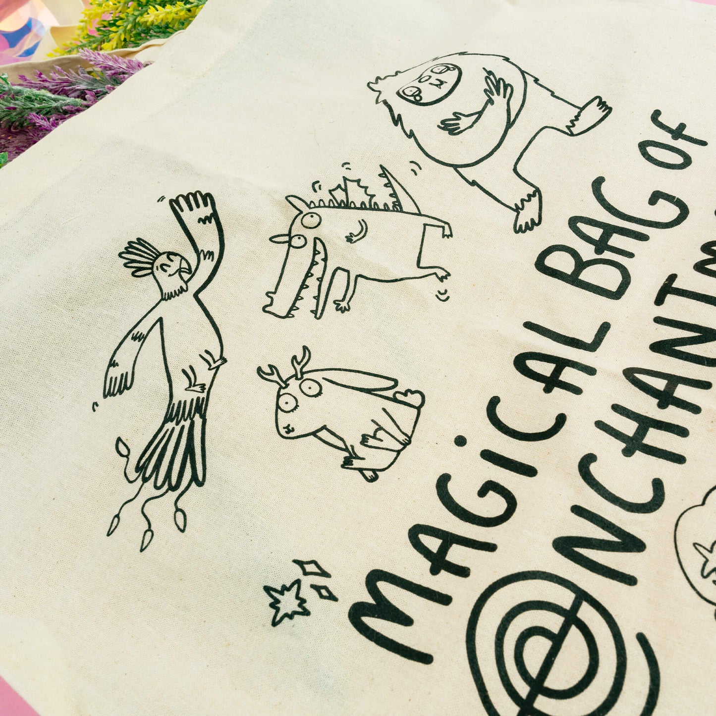 A whimsical cotton tote bag by Happiness Enchanters, titled "Magical Bag of Enchantment." The natural beige bag is illustrated with a collection of charming, hand-drawn magical creatures, including a unicorn, mermaid, mushroom fairy, friendly monsters, and a flying bird. The bag's playful and imaginative design offers a delightful, eco-friendly way to carry your essentials while adding a touch of magic to everyday errands. Set against a pastel pink background with shimmering holographic details