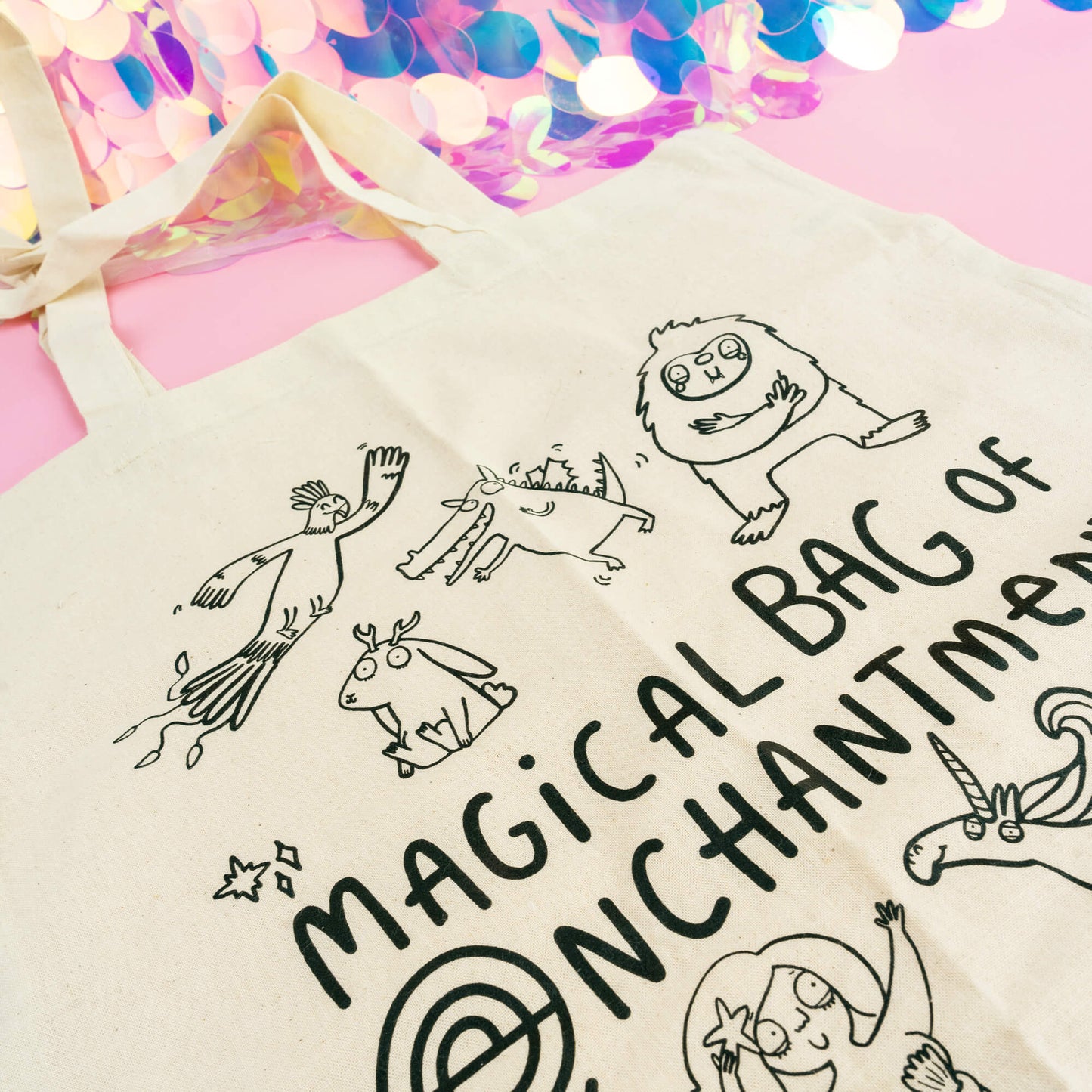 A whimsical cotton tote bag by Happiness Enchanters, titled "Magical Bag of Enchantment." The natural beige bag is illustrated with a collection of charming, hand-drawn magical creatures, including a unicorn, mermaid, mushroom fairy, friendly monsters, and a flying bird. The bag's playful and imaginative design offers a delightful, eco-friendly way to carry your essentials while adding a touch of magic to everyday errands. Set against a pastel pink background with shimmering holographic details