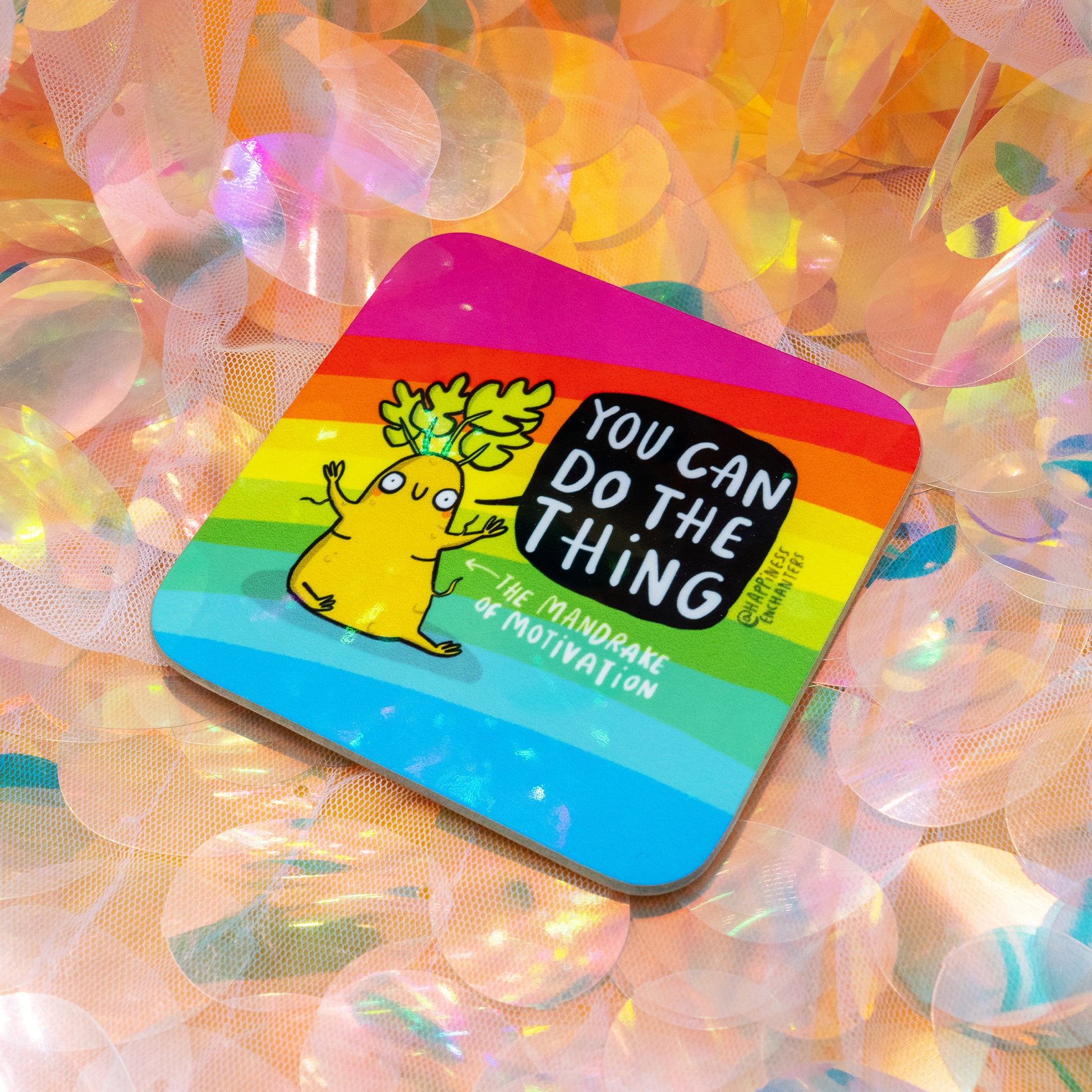 A rainbow mandrake-themed motivational coaster by Happiness Enchanters resting on iridescent sequins. The design shows a cheerful mandrake character with the message “You Can Do The Thing – The Mandrake of Motivation.” A vibrant, uplifting accessory designed by Katie Abey and Angela Sandland.