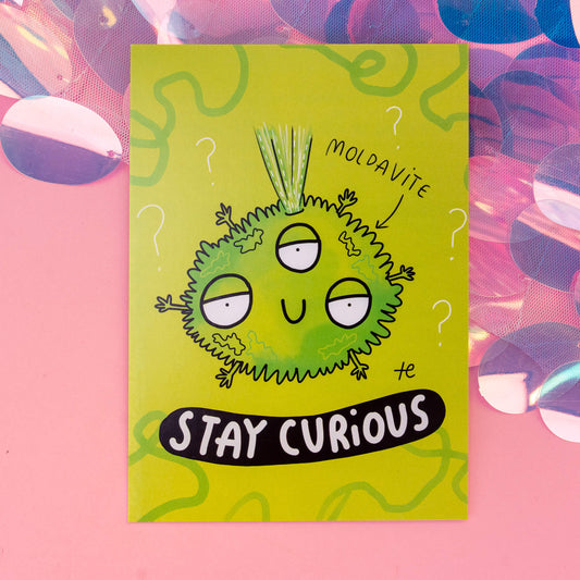 A close-up view of a Moldavite-themed postcard by 'Happiness Enchanters', displaying a charming cartoon Moldavite crystal character with three eyes and spiky hair, along with the phrase 'Stay Curious'. The background is a bright green with swirling patterns and question marks, capturing the essence of creativity and enchantment.