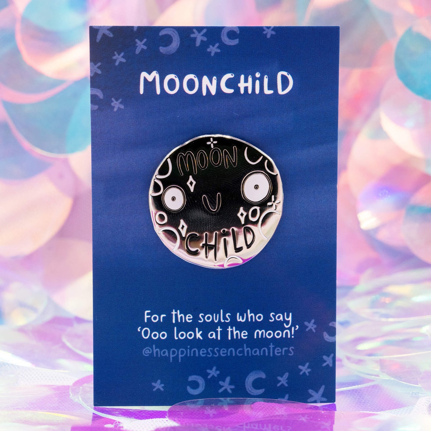 A round iridescent Moonchild pin badge by Happiness Enchanters, illustrated by Katie Abey. The brushed silver metal badge features a smiling moon face surrounded by stars and crescent moons, displayed on a deep blue backing card that reads “For the souls who say ‘Ooo look at the moon!’” against a soft pastel background.