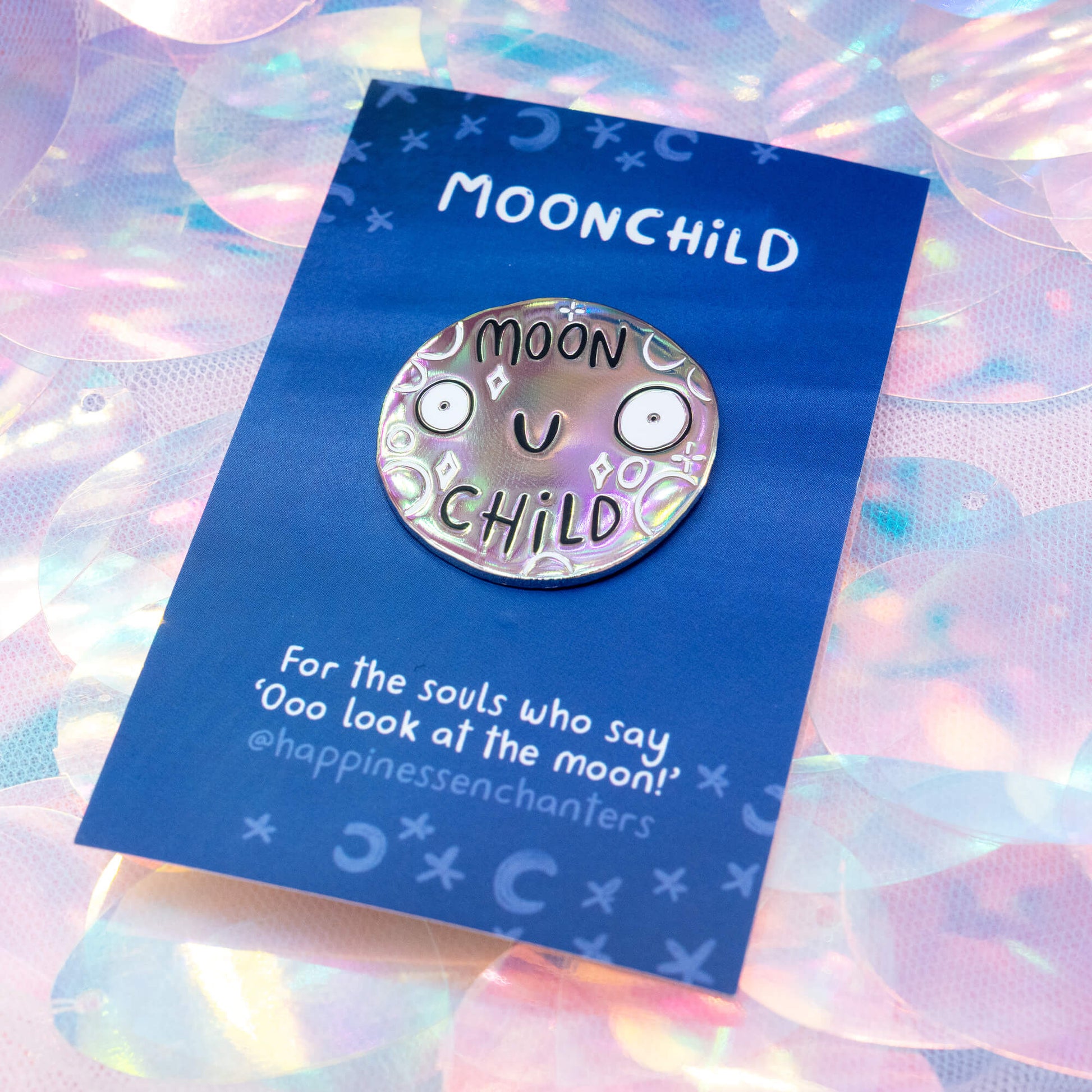 The Moonchild metal pin badge by Happiness Enchanters shown at an angle, highlighting its rainbow-toned iridescent brushed finish. The cute smiling moon face and celestial motifs shimmer softly on a blue card with the words “For the souls who say ‘Ooo look at the moon!’”.