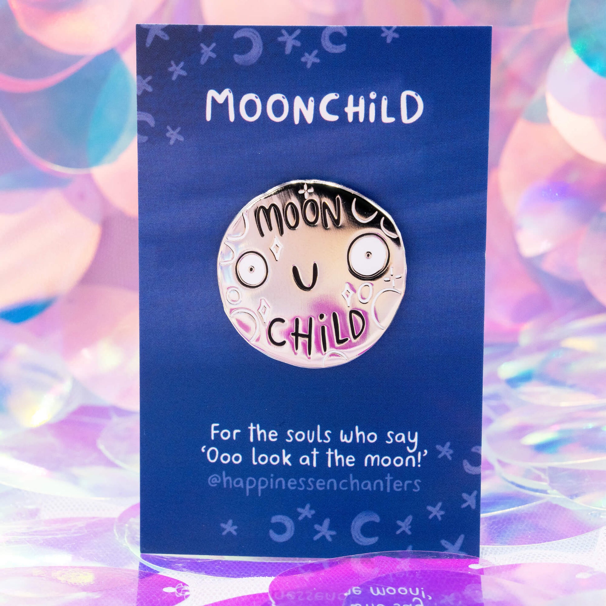 Close-up of the Moonchild enamel pin badge by Happiness Enchanters, illustrated by Katie Abey. The moon’s happy face and celestial details glow with a reflected iridescent sheen on a blue backing card. Perfect for moon lovers and dreamers who adore magical, cosmic accessories.