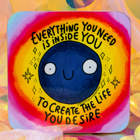 A vibrant moon coaster by Happiness Enchanters showing a happy navy blue moon in the centre of a rainbow aura. The text says “Everything You Need Is Inside You To Create The Life You Desire”. Created by Katie Abey and Angela Sandland. Photographed against shimmering iridescent sequins for a dreamy, magical look.