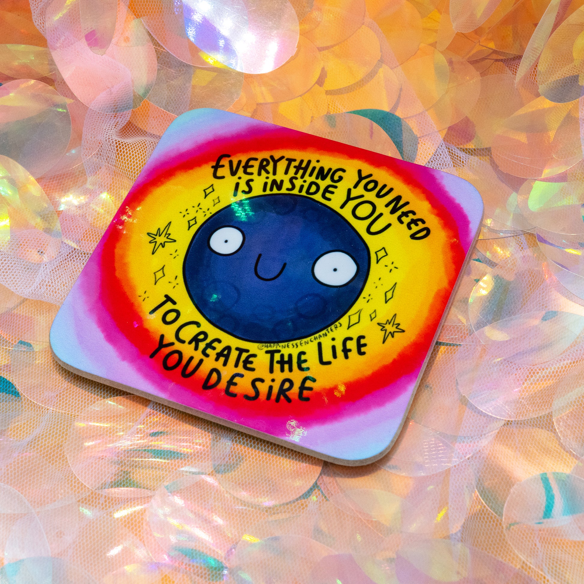 A colourful square coaster by Happiness Enchanters featuring a smiling navy blue moon surrounded by a glowing rainbow aura. The text reads “Everything You Need Is Inside You To Create The Life You Desire”. Designed by Katie Abey and Angela Sandland. The coaster rests on iridescent sequins with a soft magical vibe.