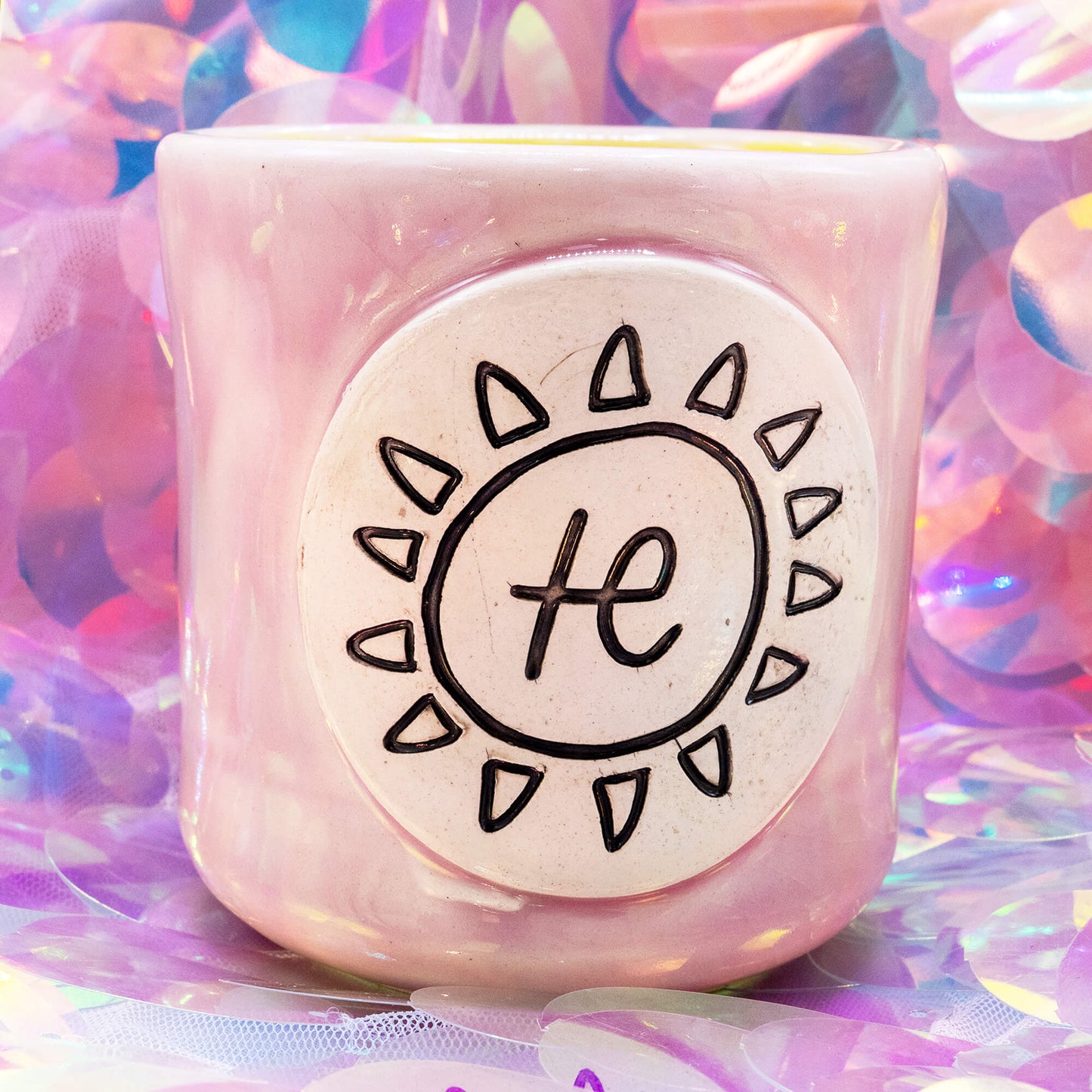 Pale pink handmade ceramic mug with the Happiness Enchanters sun and symbol design on a white medallion, shown on an iridescent pastel background.