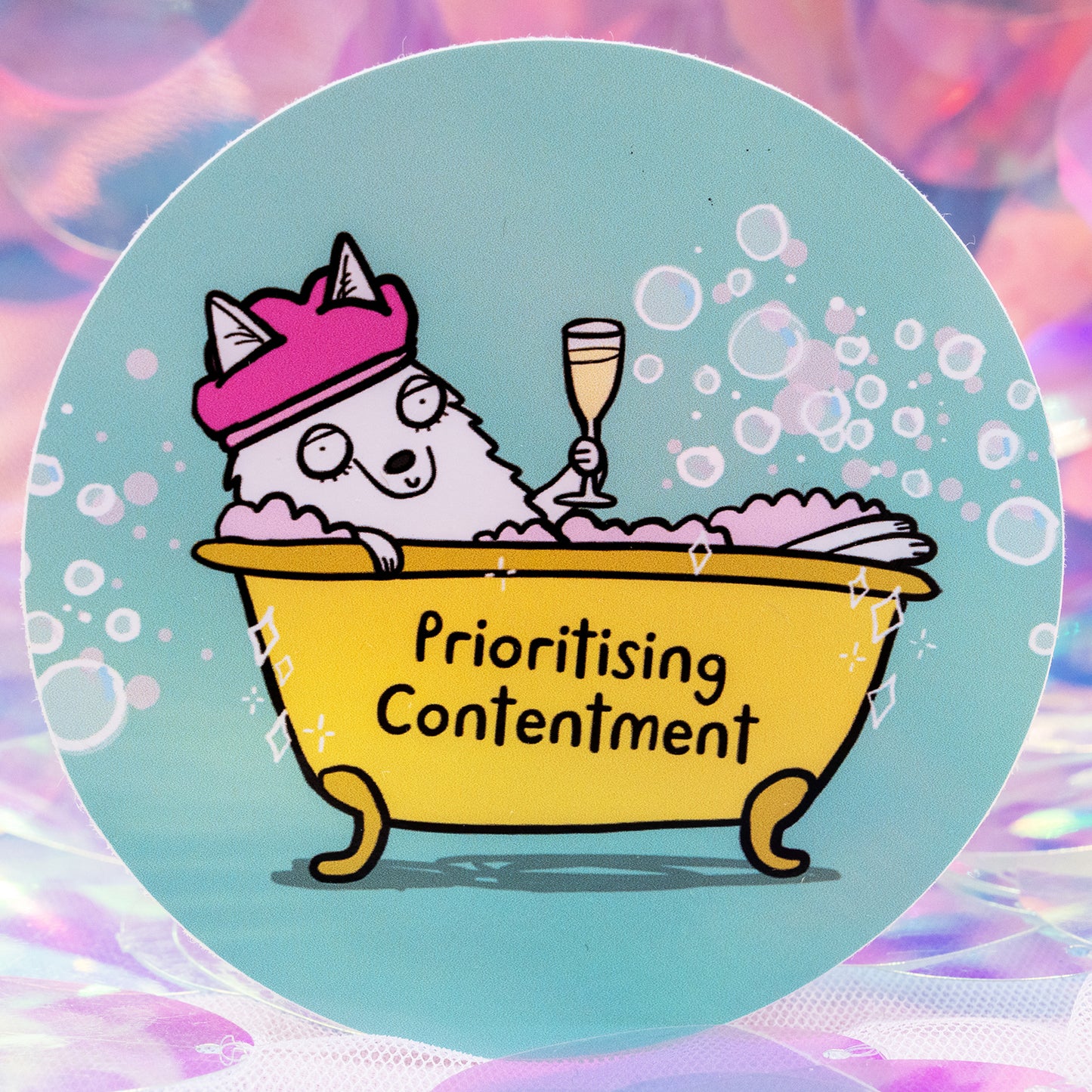 A round vinyl sticker by Happiness Enchanters featuring a cute illustrated wolf relaxing in a yellow bubble bath, wearing a pink shower cap and holding a champagne glass. The tub reads “Prioritising Contentment”. The background is soft turquoise with bubbles. Designed by Katie Abey for gentle, magical self care.