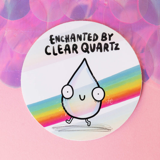  A round sticker featuring a cute clear quartz crystal character with big eyes and a smile, set on a vibrant pastel rainbow background. The words "Enchanted by Clear Quartz" are written above. The sticker is displayed on a pink surface with shimmering accents, evoking a cheerful, enchanted vibe. Designed by Happiness Enchanters, known for blending creativity with alchemy and wellness.