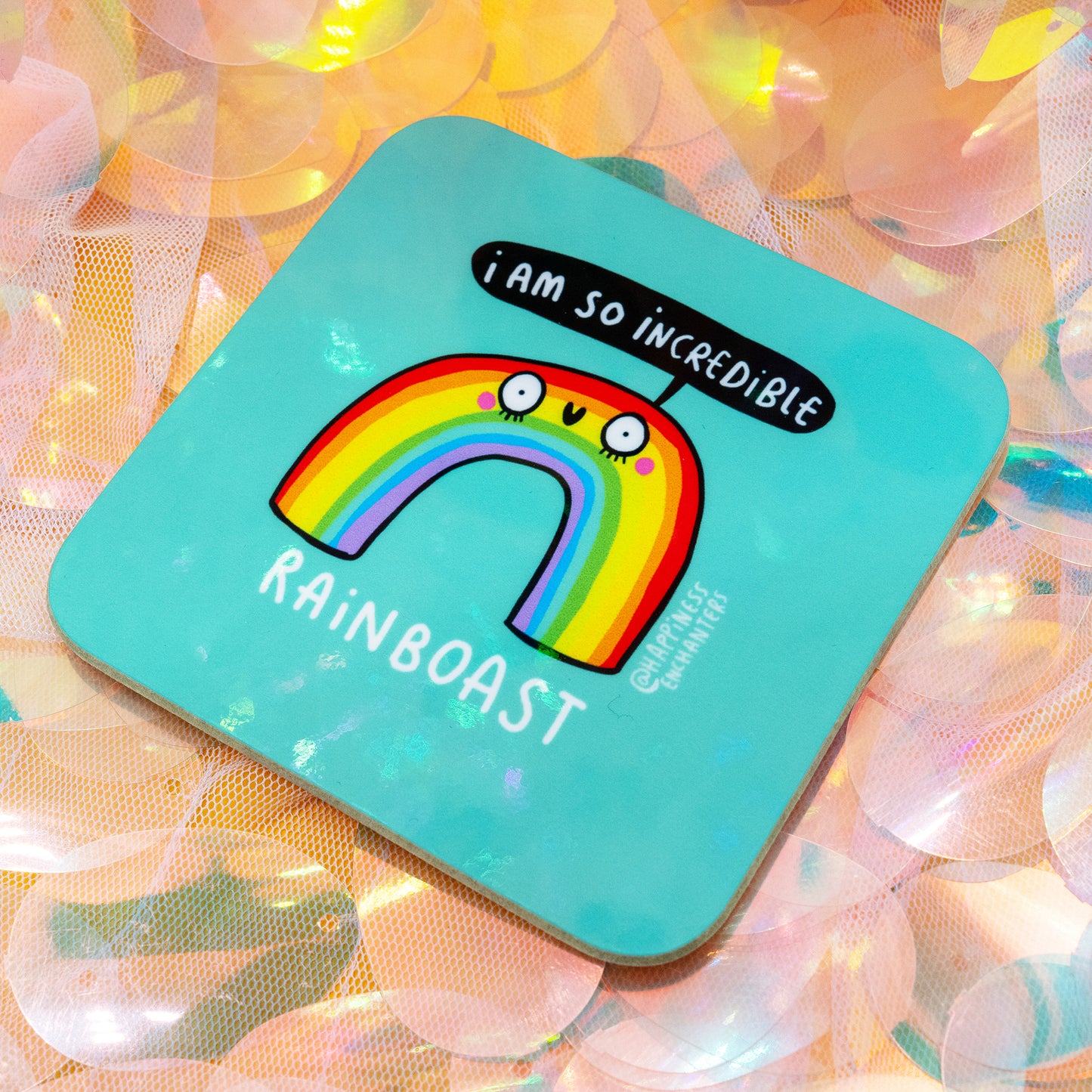 A whimsical Rainboast coaster by Happiness Enchanters displayed on pastel iridescent sequins. The teal coaster shows a bright rainbow character with a happy face and the phrase “I am so incredible.” Created by artists Katie Abey and Angela Sandland, this colourful homeware piece brings uplifting charm to your desk, kitchen, or gifting moments.