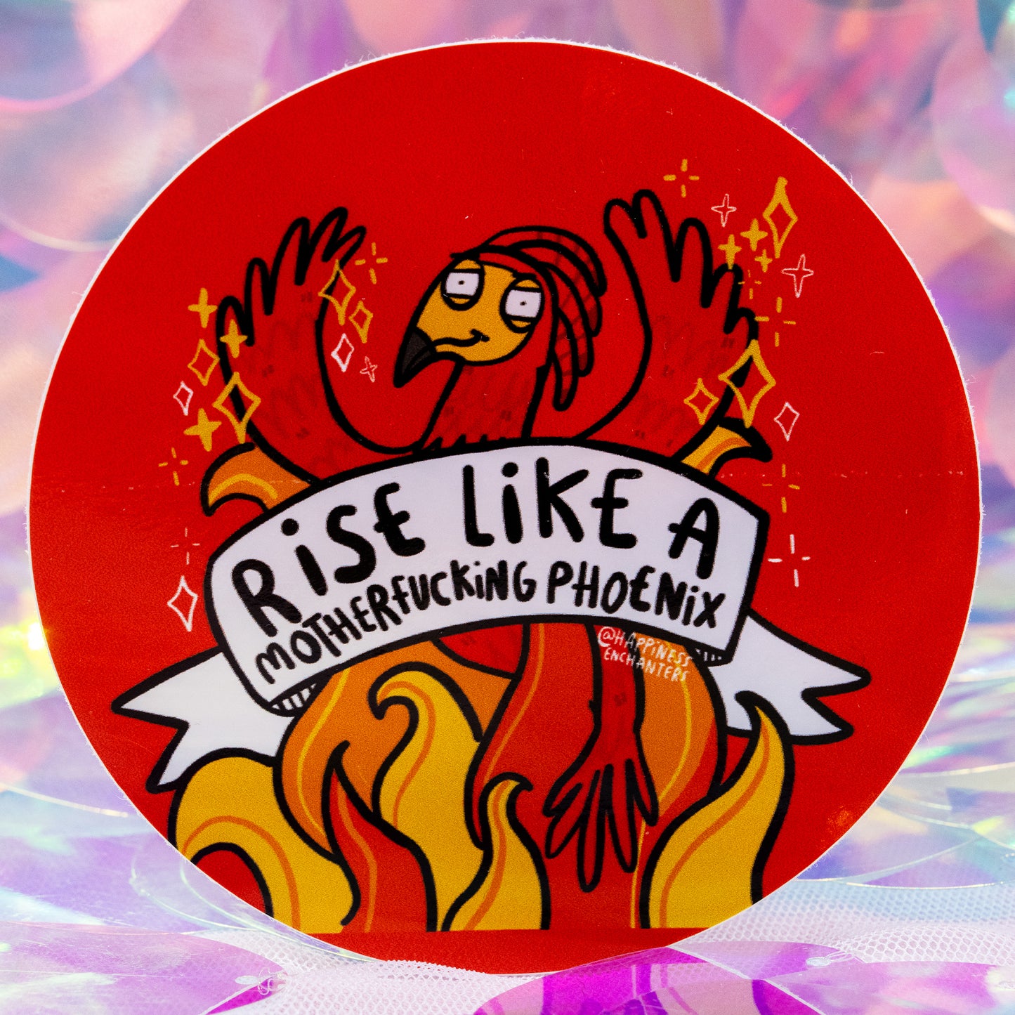 Close up of the round red “Rise Like A Motherfucking Phoenix” sticker by Happiness Enchanters. The illustrated phoenix spreads its wings with sparkles around it, rising from bright orange flames. Designed by artists Katie Abey and Angela Sandland. The background is soft, pastel and iridescent.
