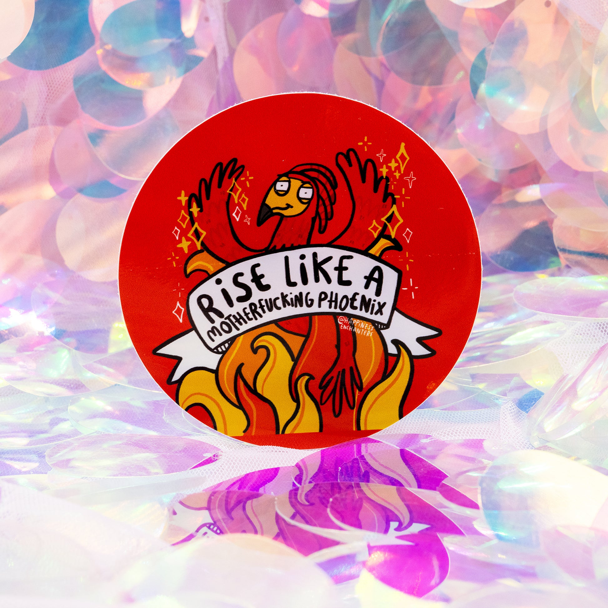 A round red sticker featuring a whimsical illustrated phoenix rising from bright orange flames. The phoenix has raised wings, sparkles around it, and a banner reading “Rise Like A Motherfucking Phoenix”. Designed by Happiness Enchanters, created by Katie Abey and Angela Sandland. The sticker sits on a magical, iridescent background.
