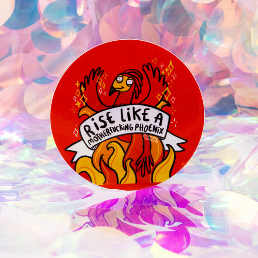 A round red sticker featuring a whimsical illustrated phoenix rising from bright orange flames. The phoenix has raised wings, sparkles around it, and a banner reading “Rise Like A Motherfucking Phoenix”. Designed by Happiness Enchanters, created by Katie Abey and Angela Sandland. The sticker sits on a magical, iridescent background.
