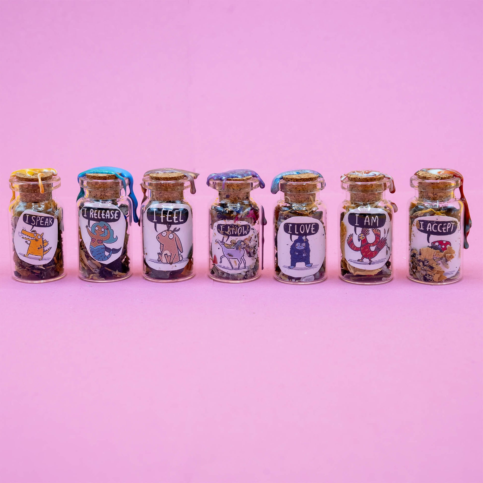 A set of seven handcrafted Word Spell bottles by Happiness Enchanters, arranged in a row on a pastel pink background. Each bottle contains a unique mix of herbs and crystals, sealed with coloured wax and corks. Illustrated labels show magical characters with affirmations: "I Speak," "I Release," "I Feel," "I Know," "I Love," "I Am," and "I Accept." Created by Katie Abey & Angela Sandland for meditation and intention setting.